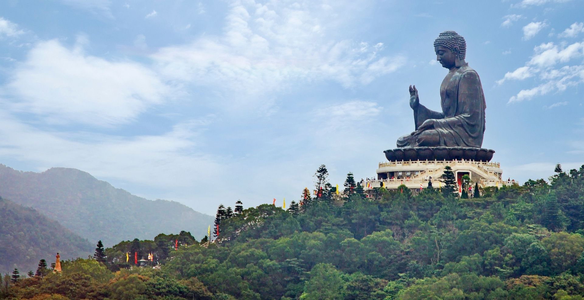 Une grande statue de Bouddha en bronze se dresse au sommet d'une colline boisée, sur une plate-forme à plusieurs niveaux, sous un ciel partiellement nuageux.