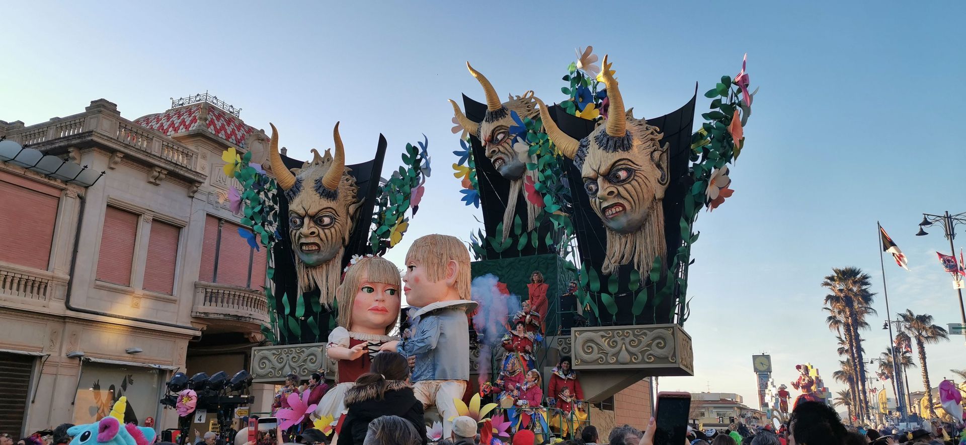 Un elaborato carro di carnevale, caratterizzato da grandi teste di demoni cornuti e due bambole giganti, attraversa una strada affollata.