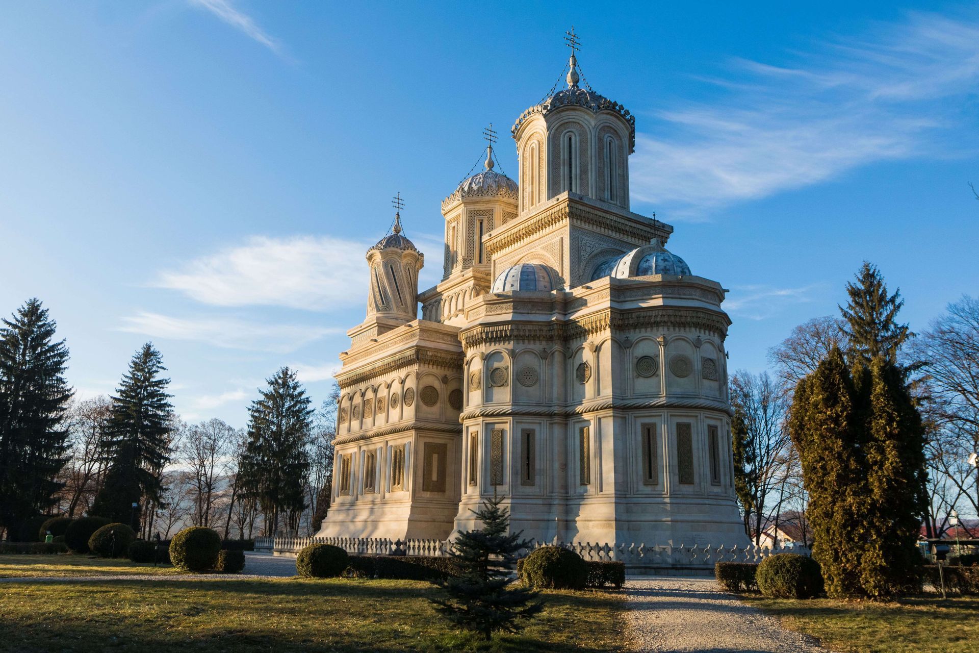 Une cathédrale ornée, de couleur claire, avec de multiples dômes et tours, se dresse dans un parc verdoyant sous un ciel bleu clair.