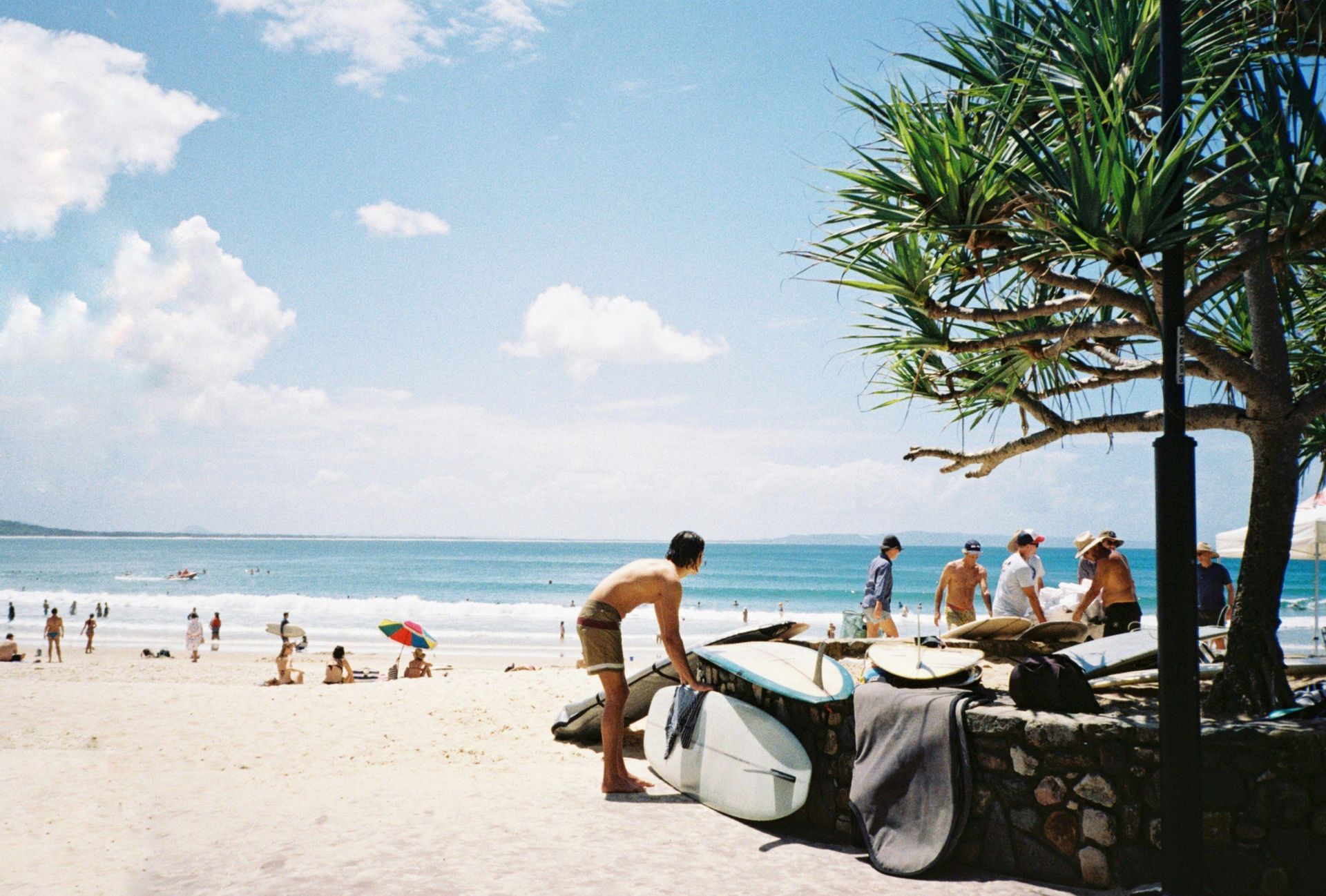 Un surfeur prépare sa planche sur un mur de pierre sur une plage animée, avec l'océan et d'autres personnes en arrière-plan.