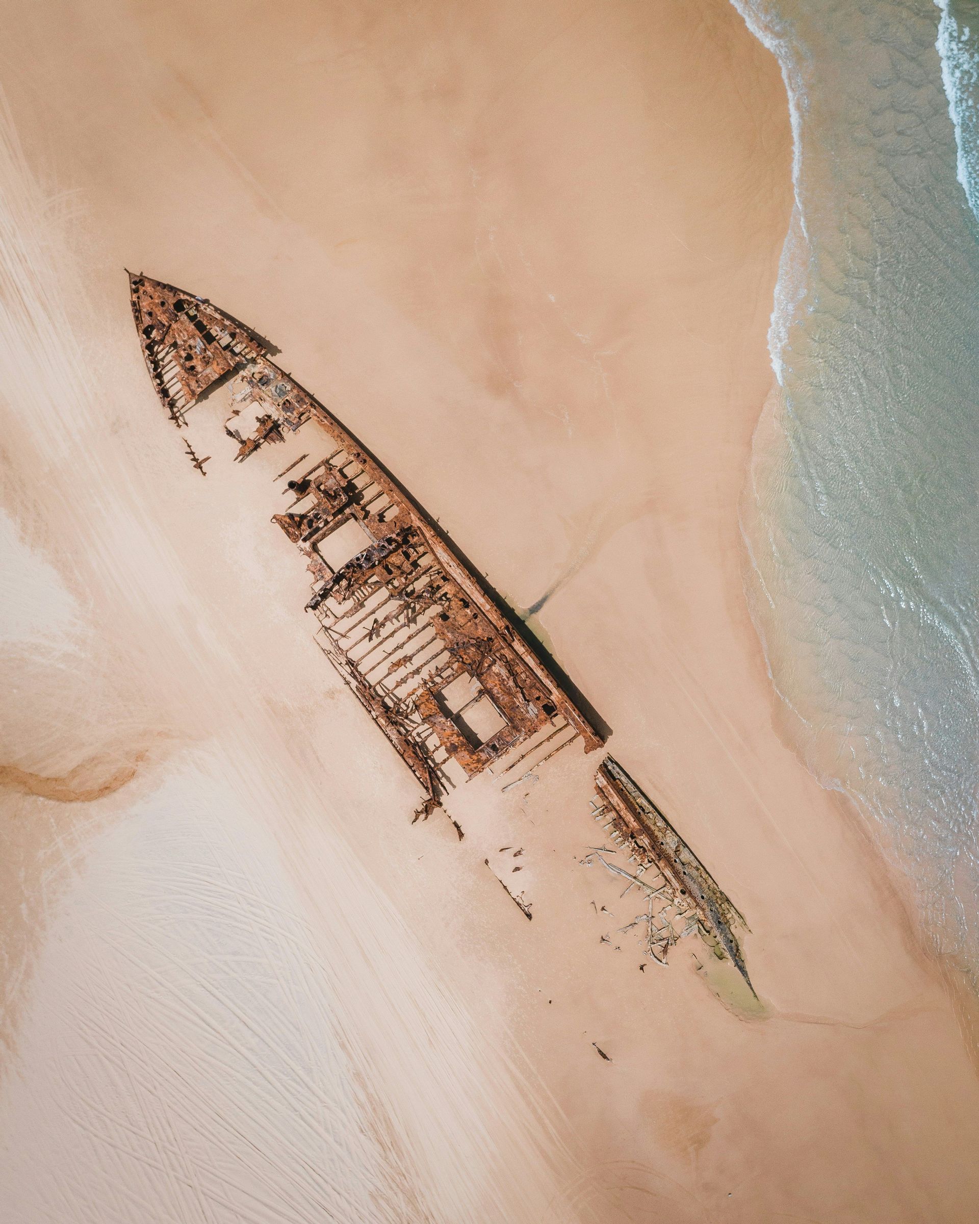 Vue aérienne d'une grande épave rouillée échouée sur le sable, avec de douces vagues de l'océan qui viennent s'échouer.