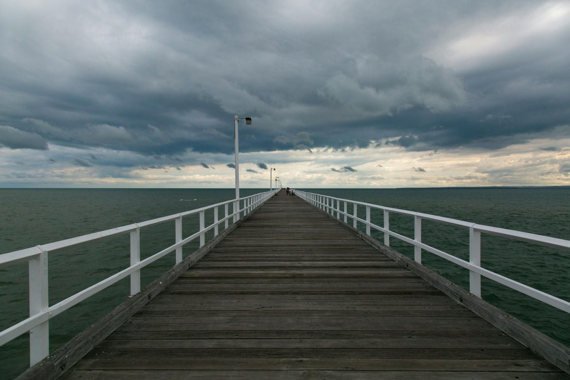 Une longue jetée en bois avec des balustrades blanches s'étend sur une mer sombre sous un ciel couvert et nuageux.