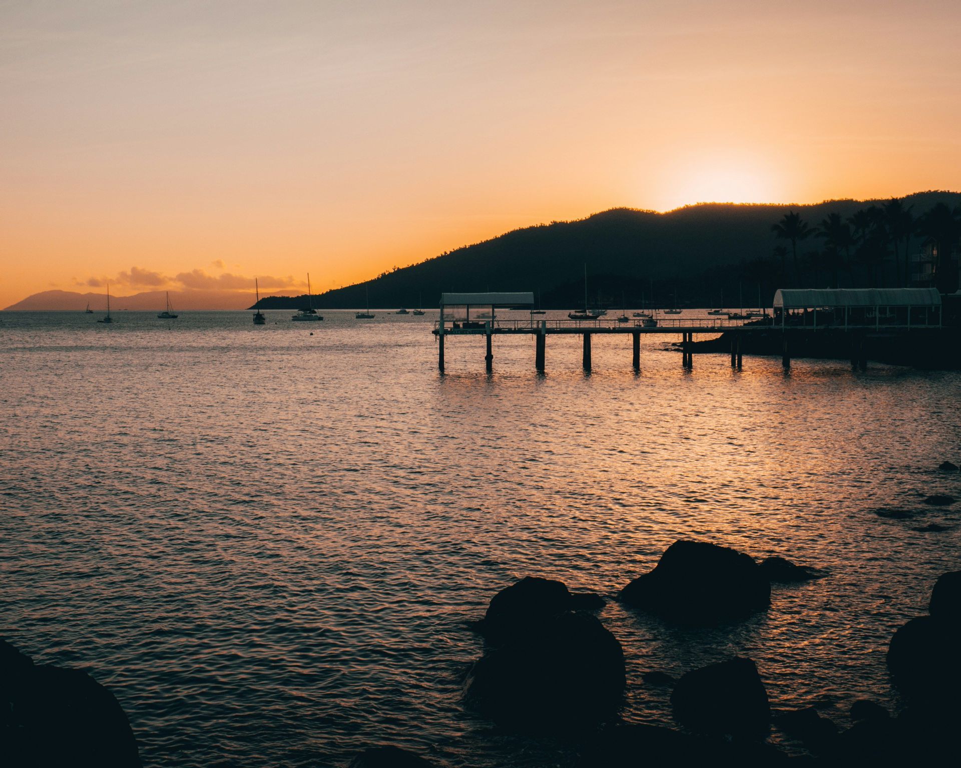Le soleil se couche derrière une colline en silhouette, projetant une lueur orange sur une baie avec une jetée et des voiliers ancrés.