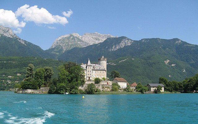 Un castello di pietra si erge su un'isola boscosa in un lago turchese, con grandi montagne coperte di foreste sullo sfondo.