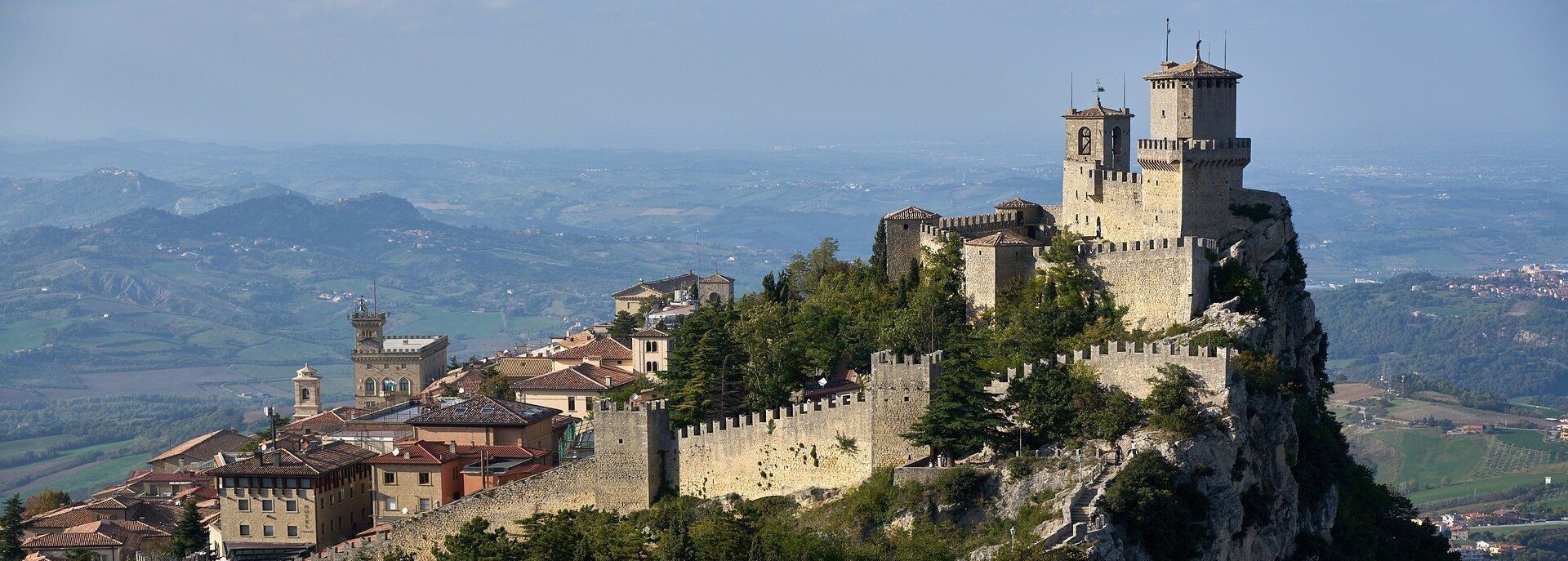 Un castello medievale in pietra con mura fortificate è arroccato su una montagna rocciosa e alberata, che domina una valle di dolci colline.