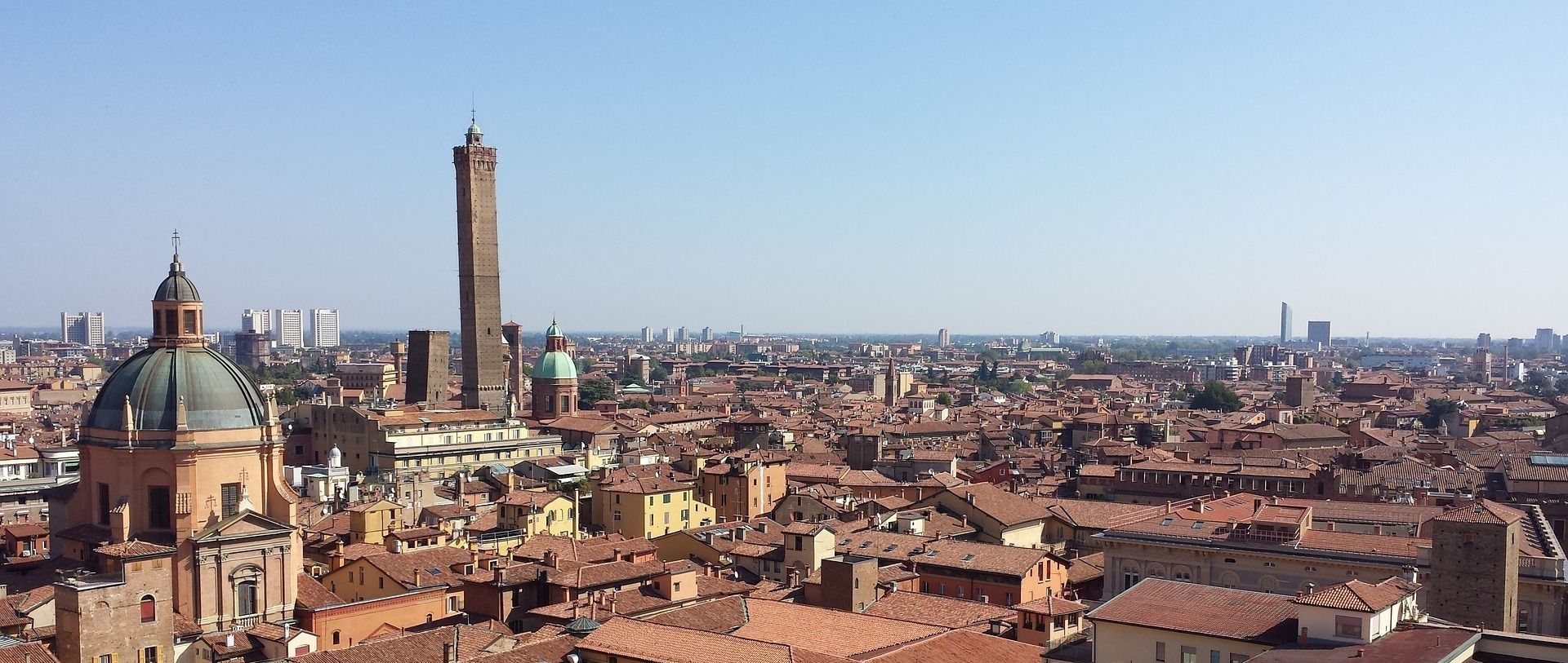 Un paesaggio urbano di tetti in terracotta, con un'alta torre di mattoni e una chiesa a cupola che si ergono contro un cielo azzurro chiaro.