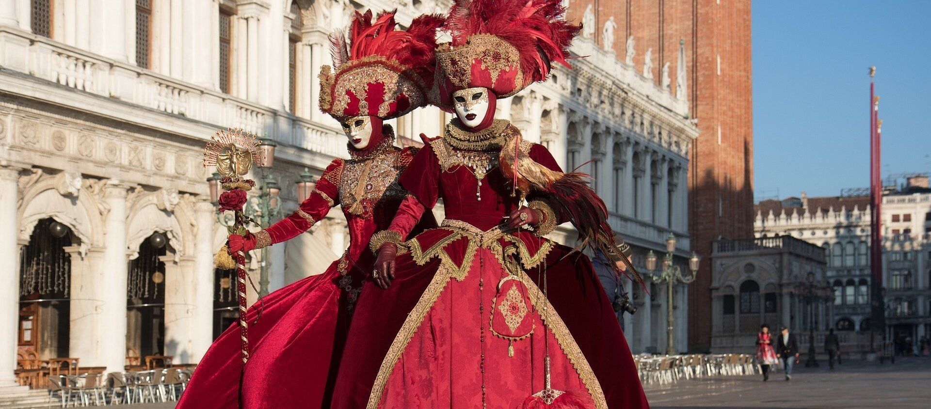 Due figure in elaborati costumi di velluto rosso, copricapi piumati e maschere bianche posano in un'ampia piazza pubblica illuminata dal sole.
