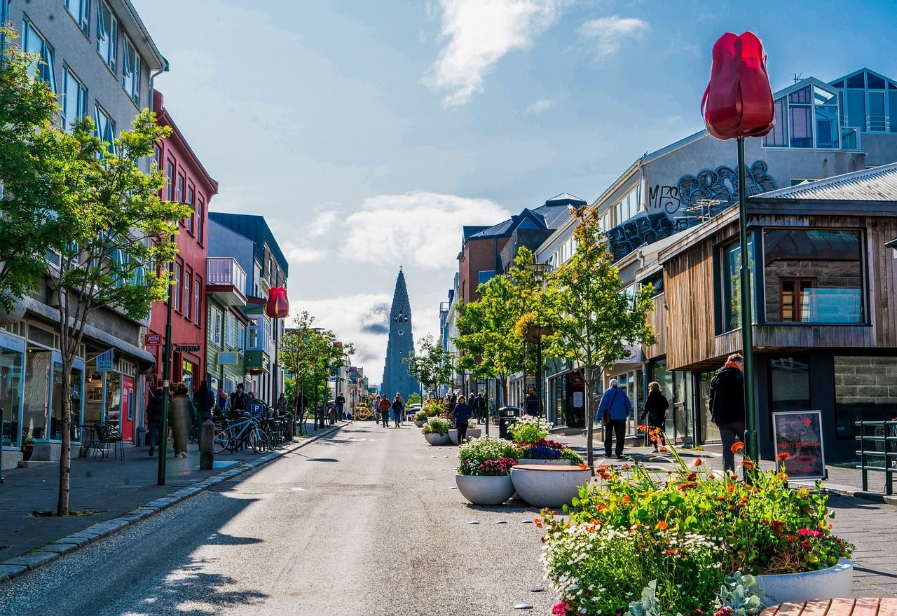 Eine sonnige Stadtstraße mit farbenfrohen Gebäuden und Bäumen, Menschen gehen auf einen hohen Kirchturm in der Ferne zu.