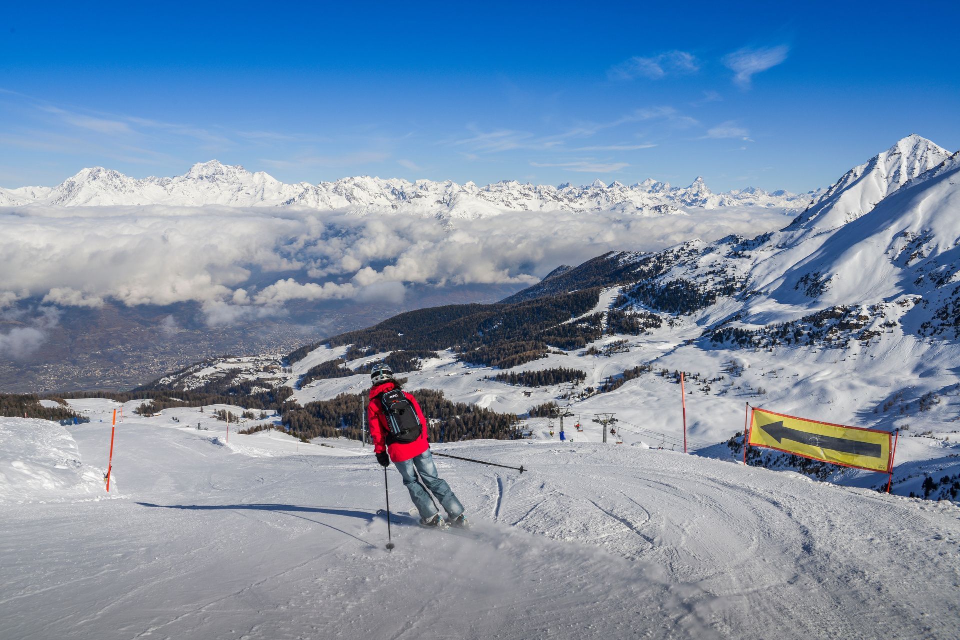Ein Skifahrer in roter Jacke fährt einen schneebedeckten Berghang hinab, mit Blick auf ein Wolkenmeer und entfernte Gipfel unter einem klaren blauen Himmel.