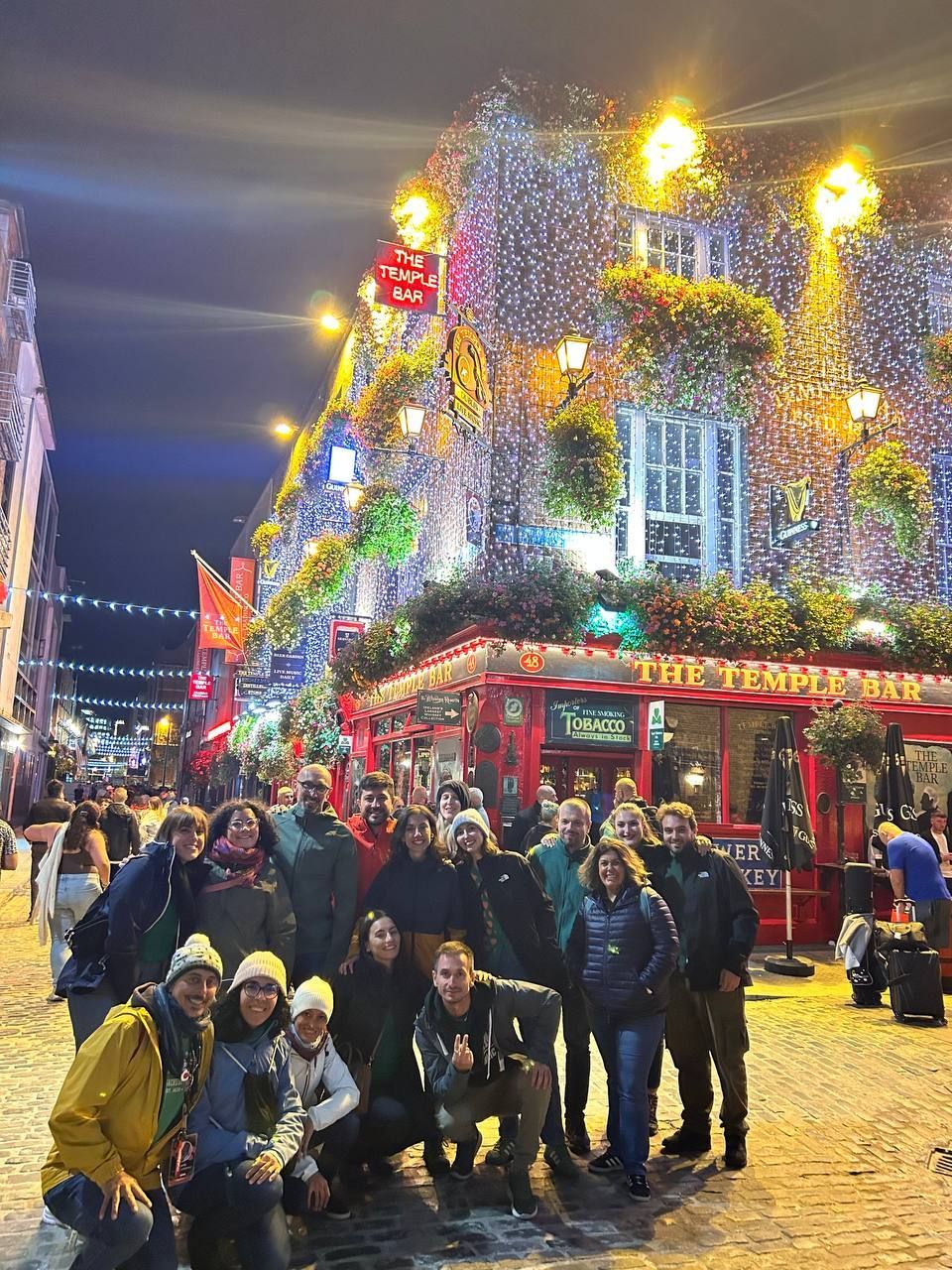 Un grupo de WeRoad posa para una foto de noche en una calle adoquinada frente a The Temple Bar, que está cubierto de luces festivas.