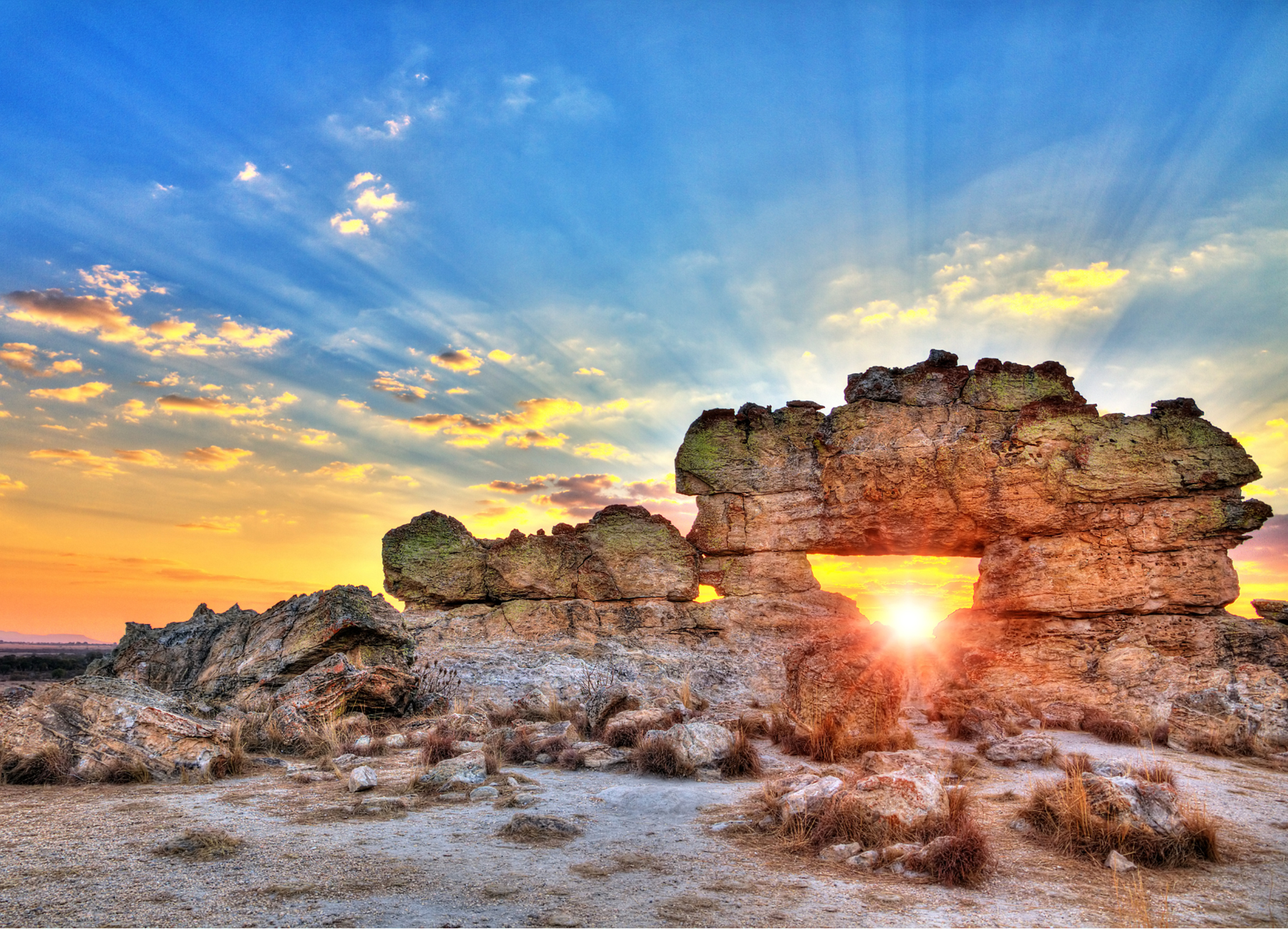 The setting sun shines through a natural arch in a large rock formation, with sunbeams radiating across a blue and yellow sky.