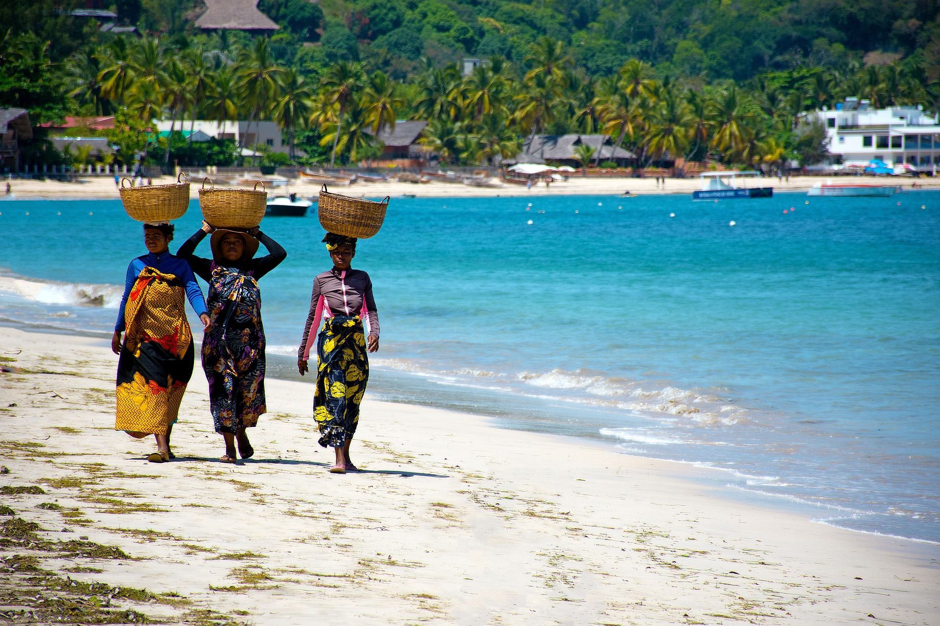 Three women in colorful clothing walk along a sandy beach, each carrying a wicker basket on her head next to turquoise water.