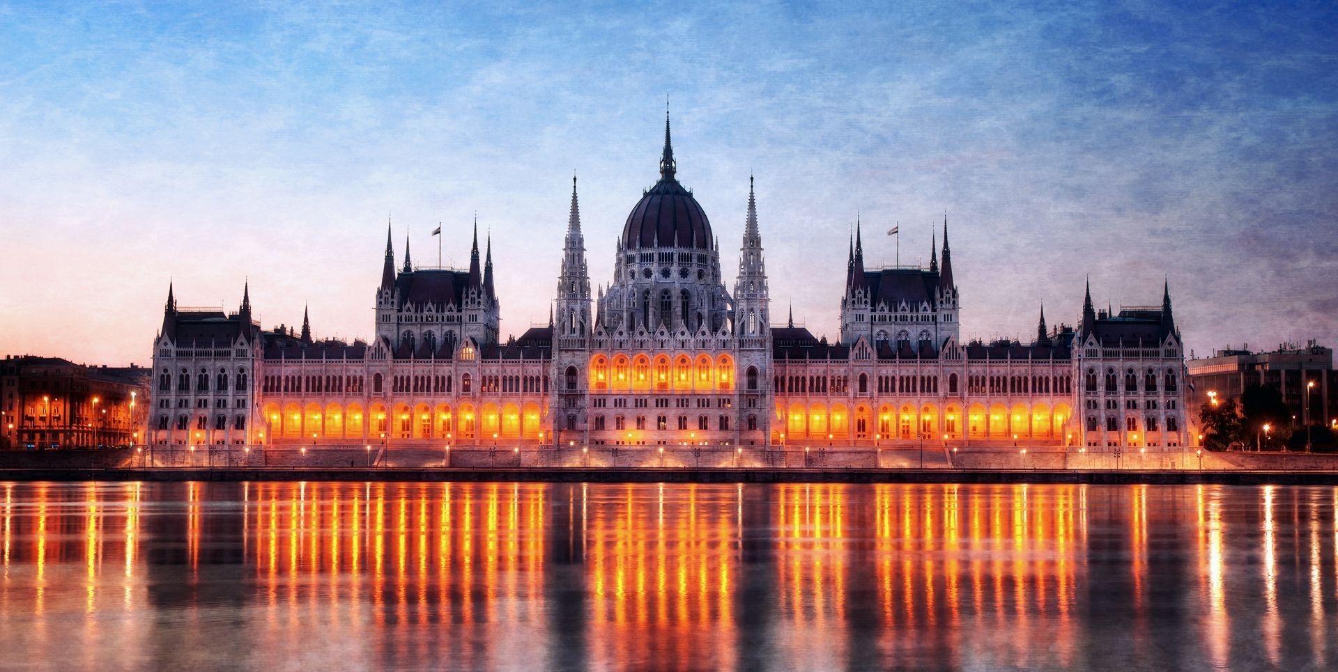 A large, illuminated government building with spires and a dome reflects in a river at twilight.