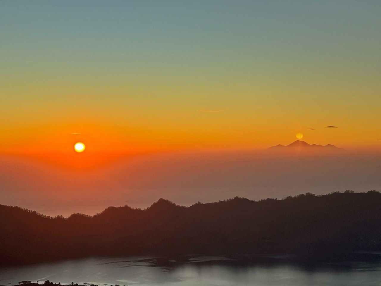 Una vivace alba crea un cielo sfumato su silhouette montuose stratificate e nebbia, con un secondo sole che emerge da una cima distante.