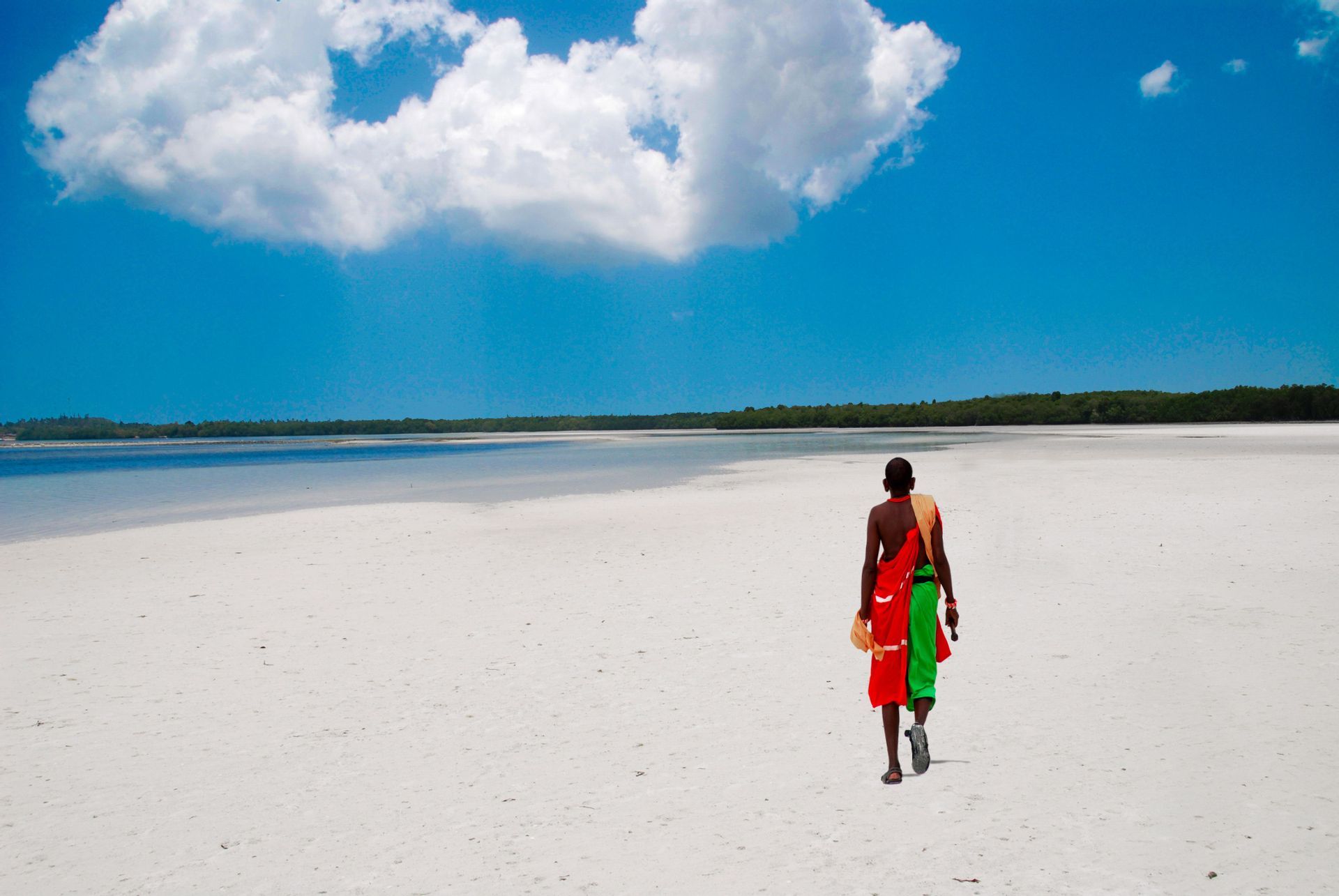 Eine Person in farbenfroher traditioneller Kleidung geht über einen weiten weißen Sandstrand unter einem blauen Himmel mit flauschigen Wolken.