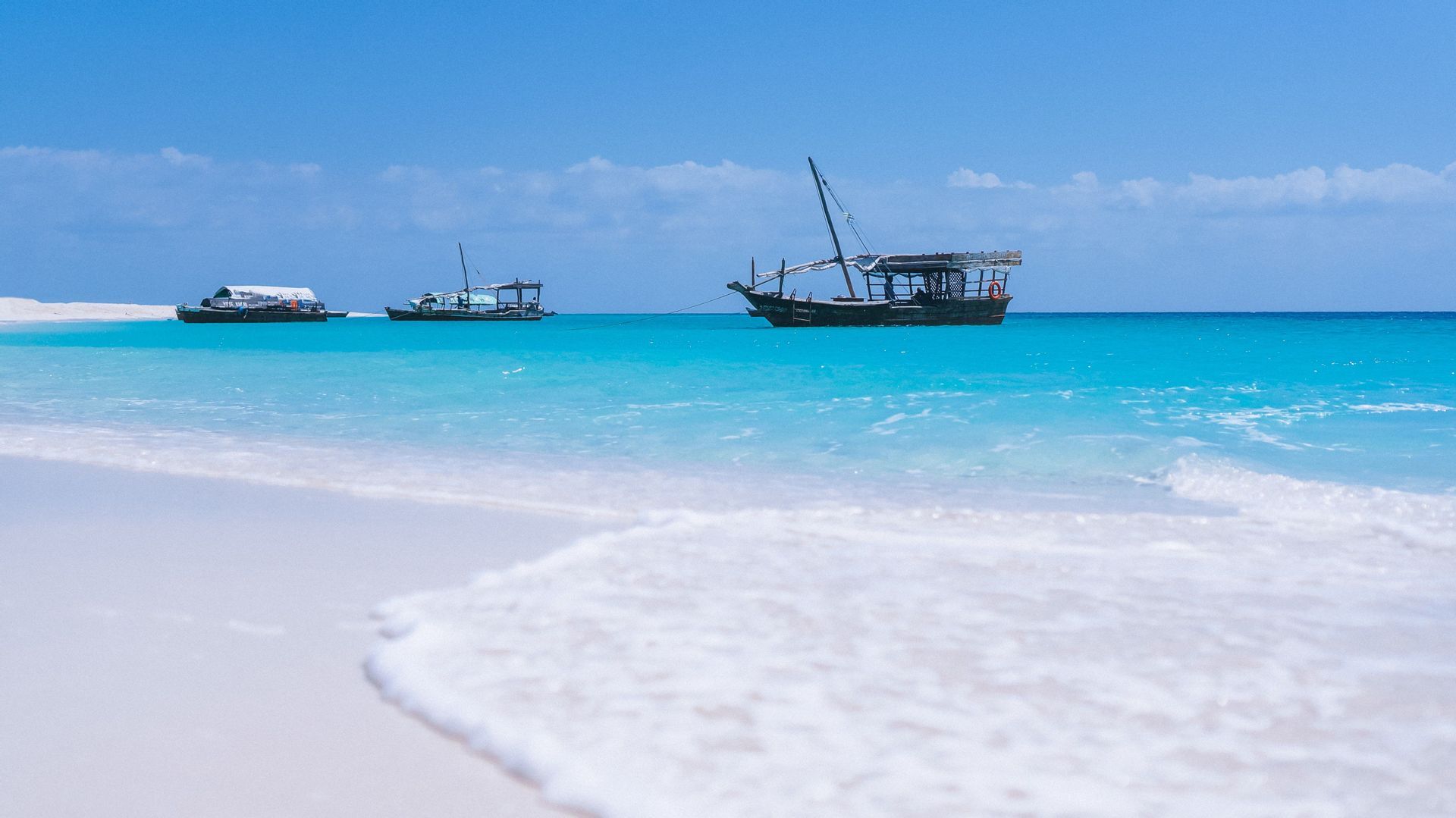 Trois bateaux en bois traditionnels ancrés dans l'eau turquoise, vus depuis une plage de sable blanc sous un ciel bleu clair.