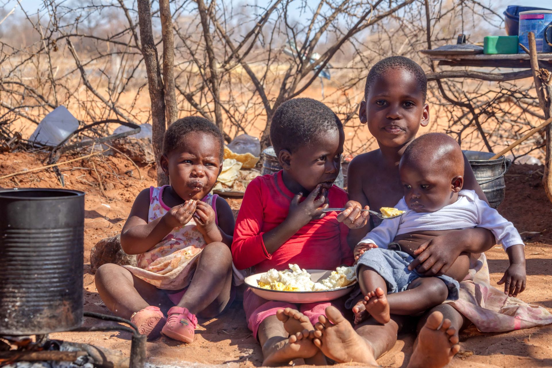 Quatre jeunes enfants sont assis par terre en extérieur et partagent un repas dans une seule assiette, l'un d'eux nourrissant un bébé.