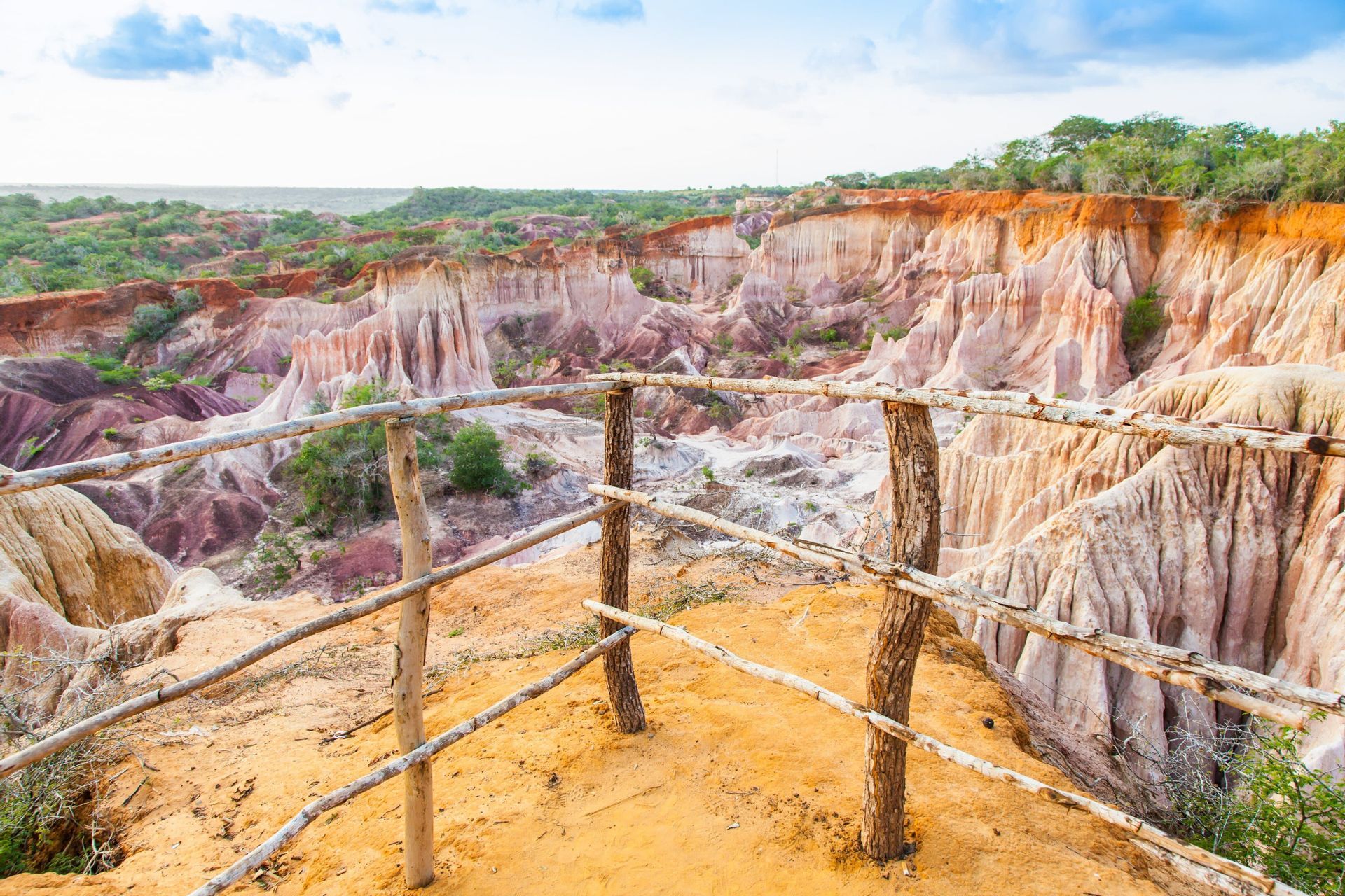 Una ringhiera in legno in un punto panoramico che si affaccia su un canyon con formazioni rocciose multicolori ed erose e alberi verdi sotto un cielo blu.