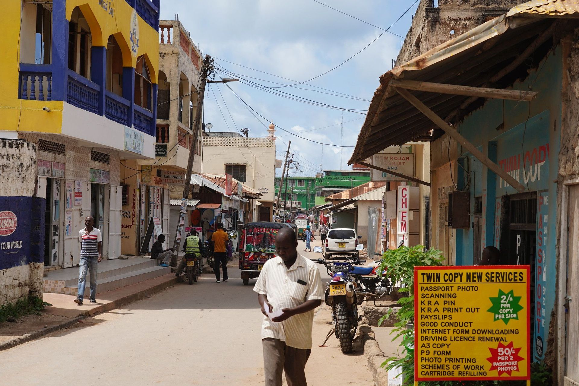 A man walks down a busy street lined with colorful buildings, shops, and motorcycles under a partly cloudy sky.