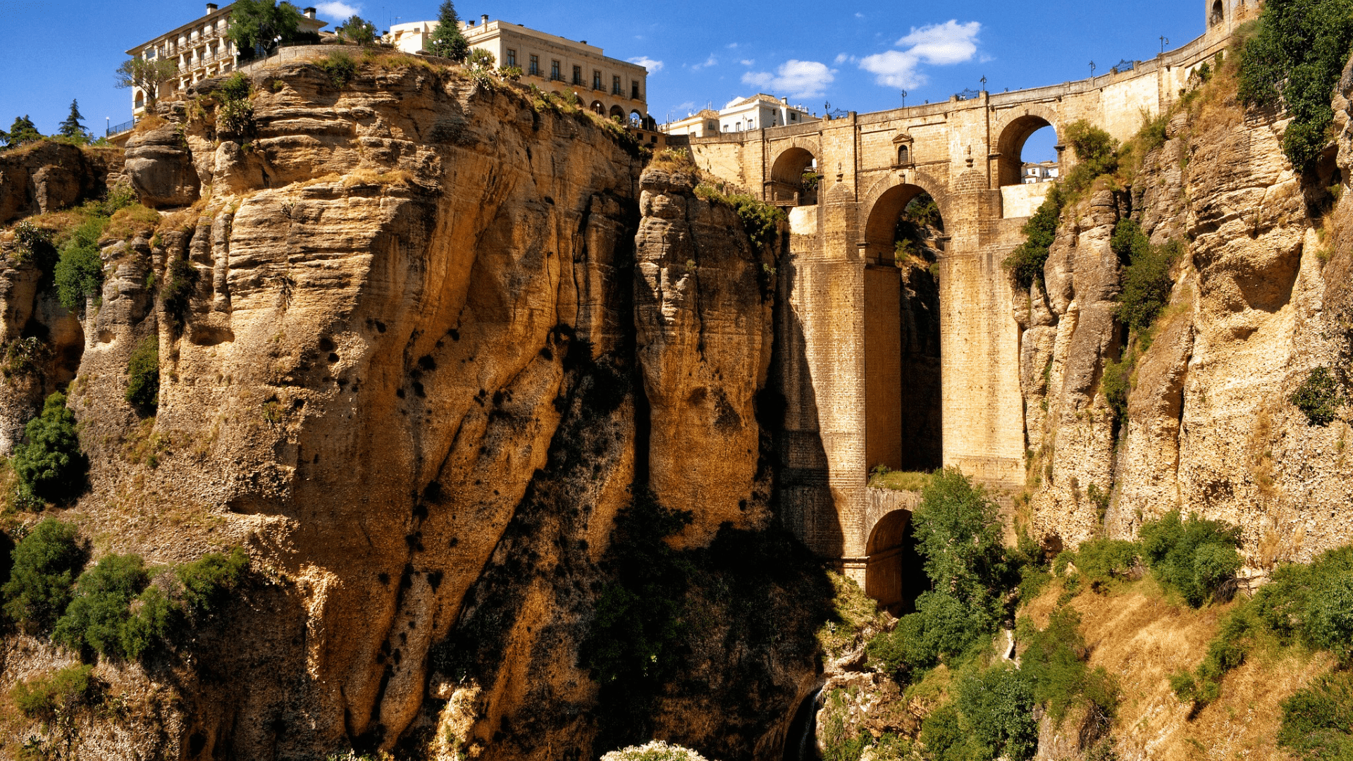 A multi-arched stone bridge connects two sheer, rocky cliffs with buildings on top under a blue sky.