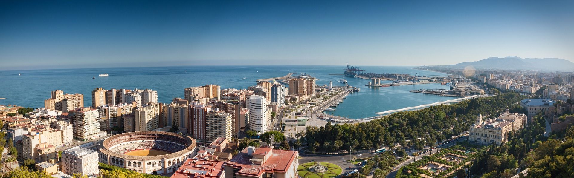 Vista aérea panorámica de una ciudad costera, destacando una plaza de toros, un gran puerto y el mar bajo un cielo azul claro.