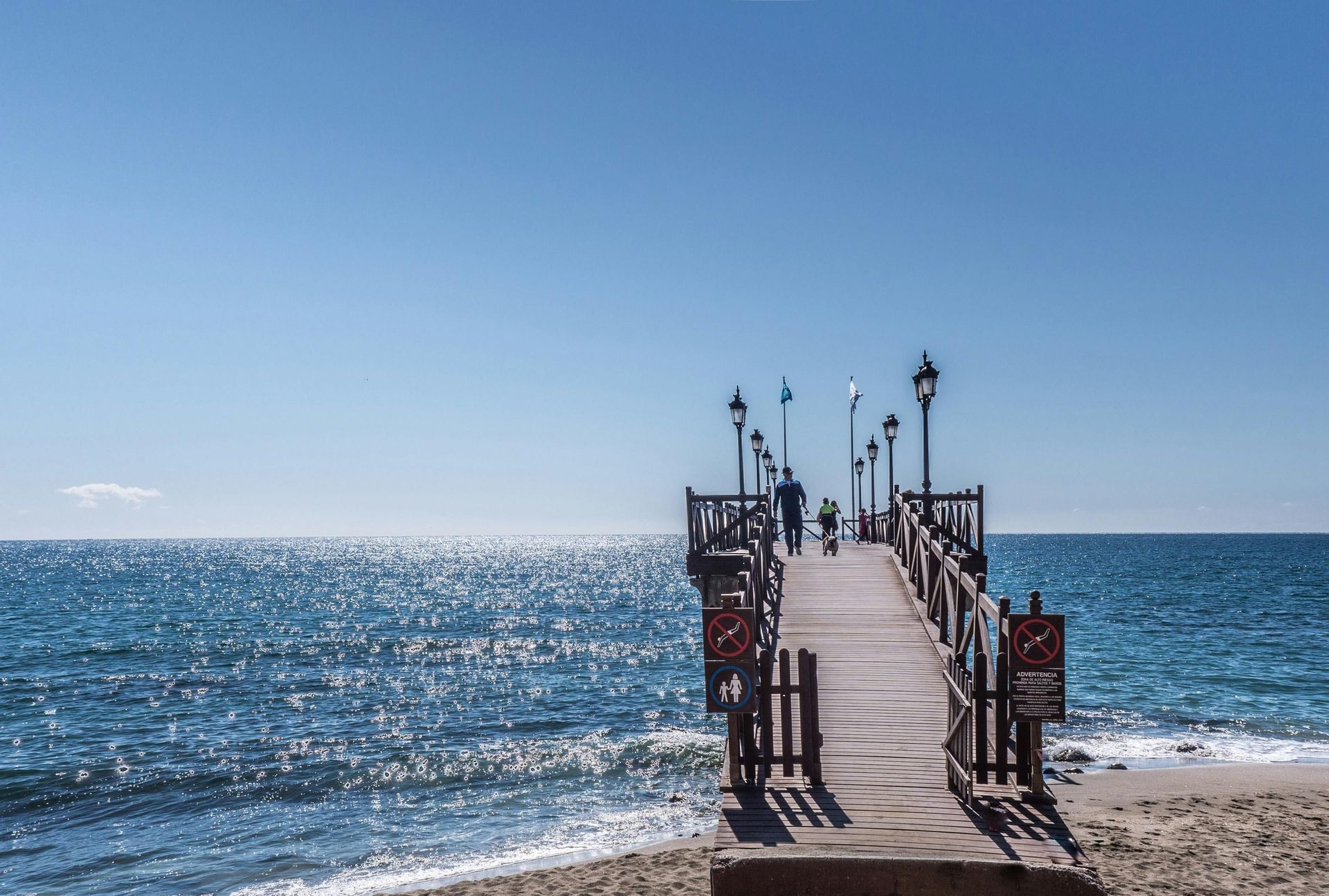 Two people and a dog walk on a wooden pier that extends from a sandy beach over the sparkling ocean.