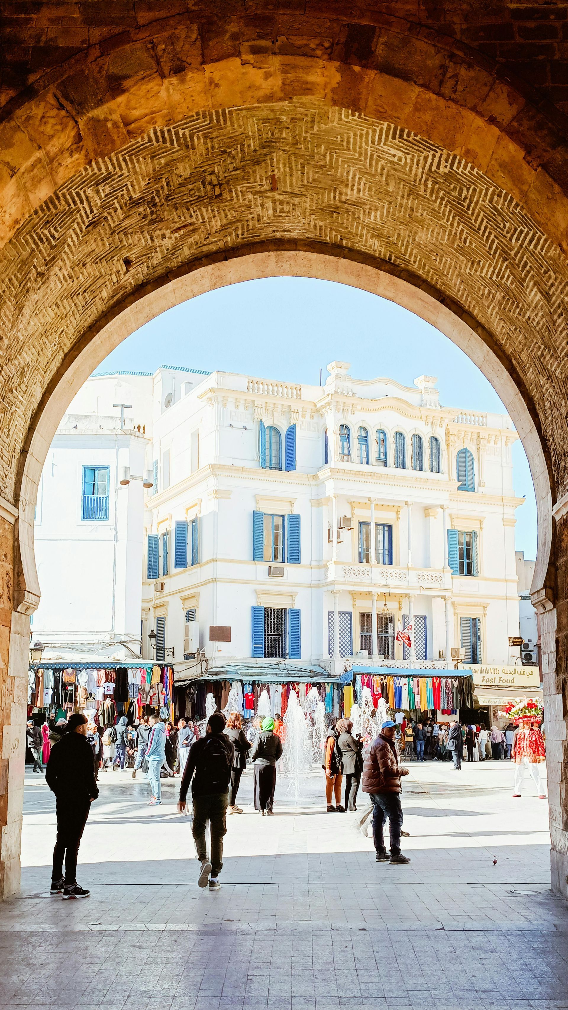 Una vista attraverso un grande arco di pietra di una soleggiata piazza pubblica con una fontana, bancarelle del mercato e un edificio bianco con persiane blu.