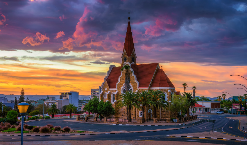 Una chiesa in pietra con tetto rosso e campanile su uno spartitraffico cittadino sotto un cielo drammatico al tramonto viola e arancione.
