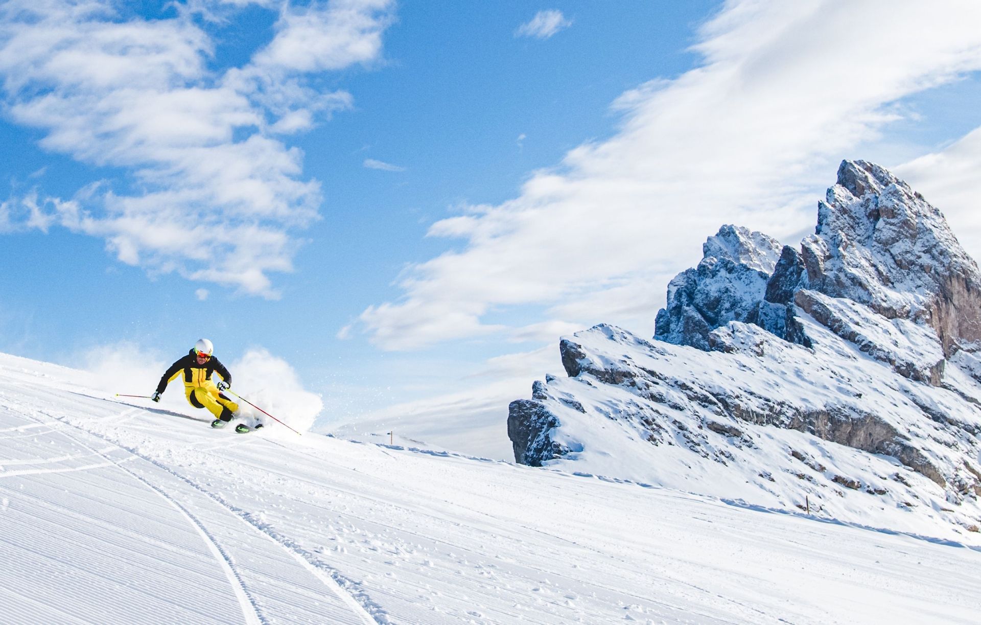 Uno sciatore in tuta gialla e nera scende una pista innevata, con cime montuose frastagliate sullo sfondo sotto un cielo parzialmente nuvoloso.