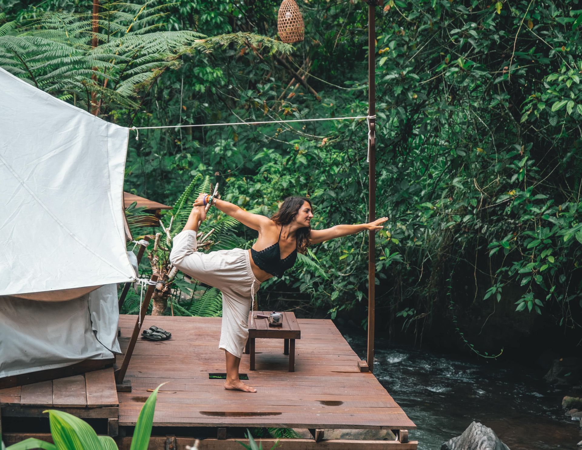 Una donna esegue una posa yoga in piedi su una piattaforma di legno accanto a un fiume, circondata da una fitta vegetazione verdeggiante della giungla.