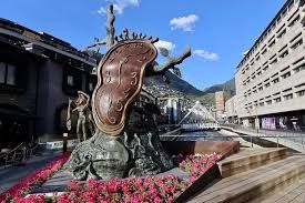 Una grande scultura di bronzo di un orologio che si scioglie si trova in una piazza cittadina, circondata da fiori rosa ed edifici moderni.