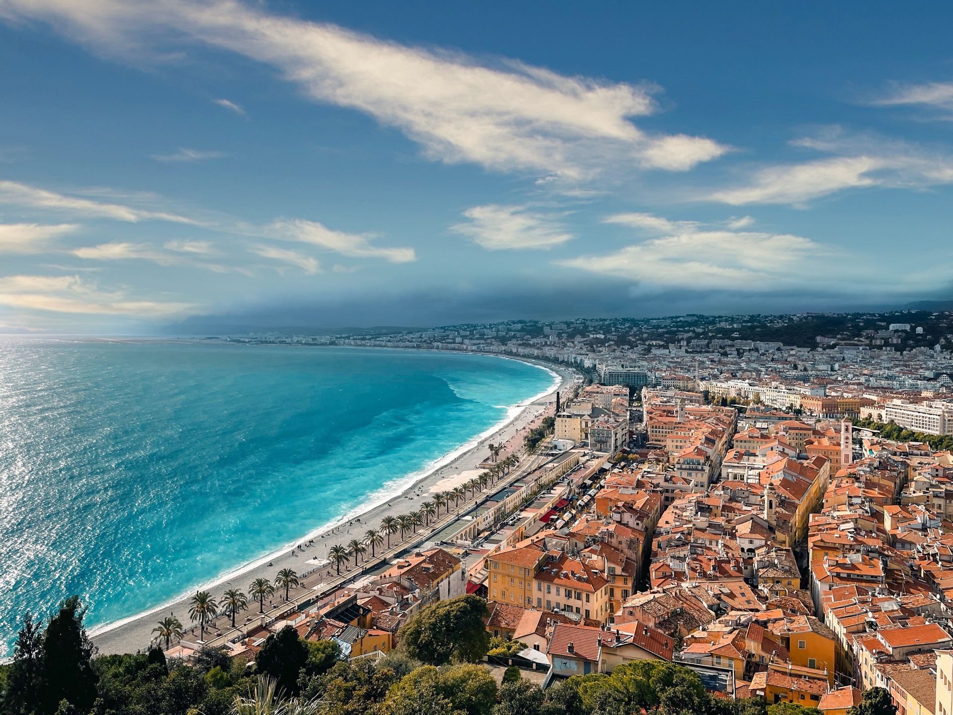Vue aérienne d'une ville côtière aux toits orangés, s'étirant le long d'une plage bordée par des eaux turquoise, sous un ciel bleu parsemé de nuages.
