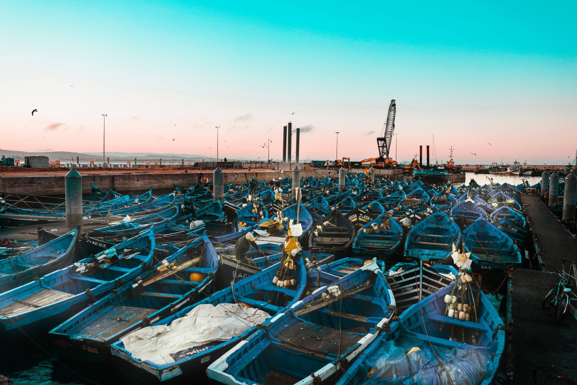 Une flotte de petits bateaux de pêche bleus est amarrée dans un port animé sous un ciel rose et turquoise.