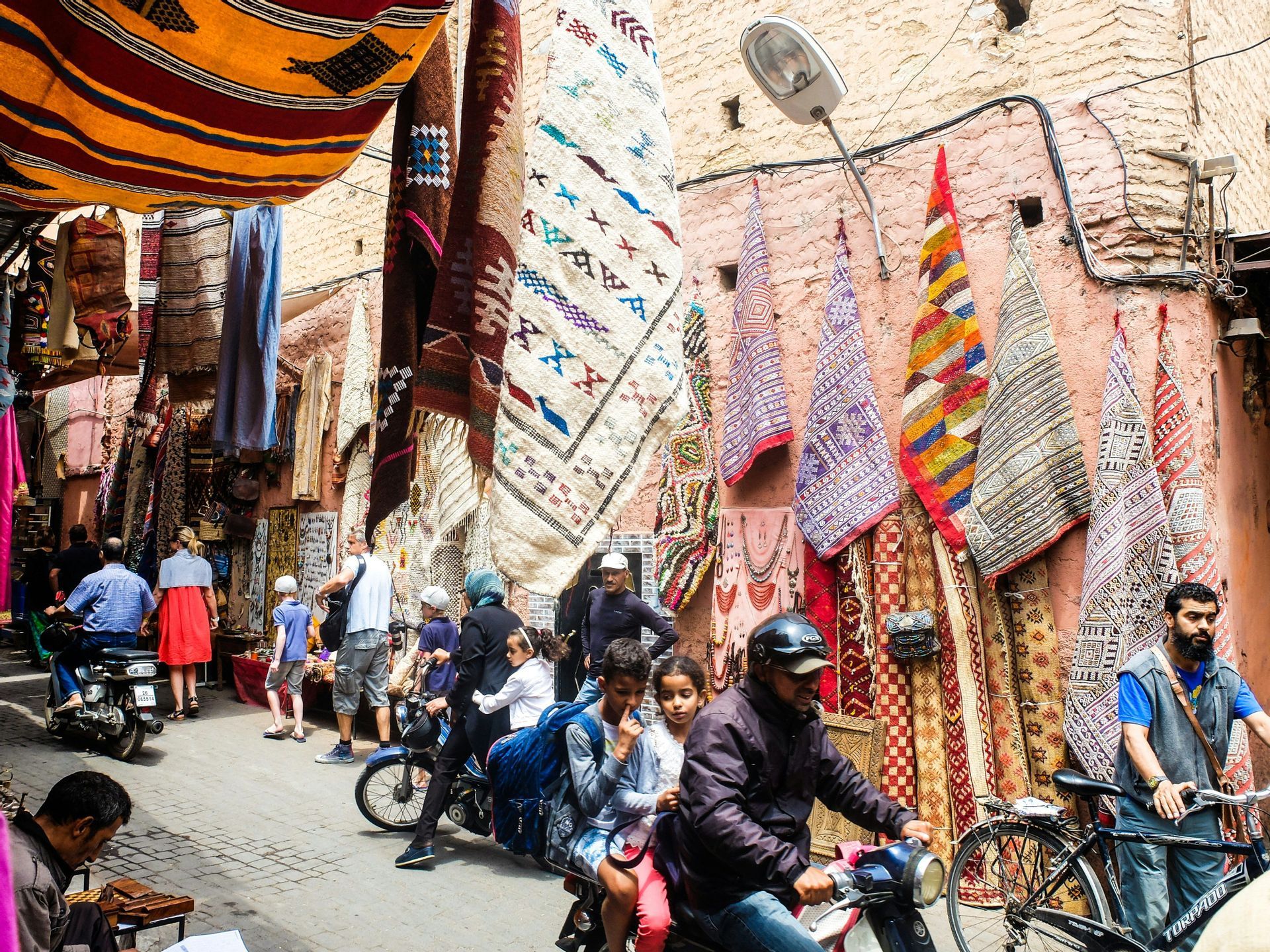 Des gens marchent et circulent à moto dans une rue de marché étroite et bondée, où des tapis et des textiles colorés sont suspendus aux bâtiments.