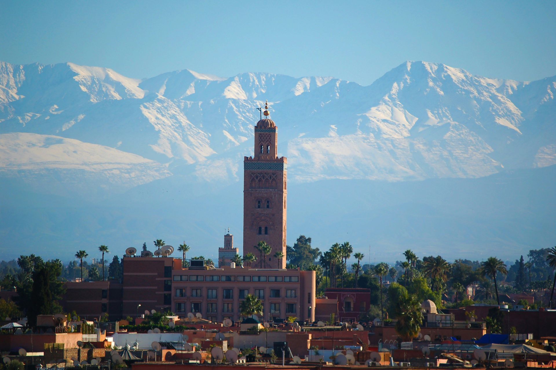 Un grand minaret brun-rouge s'élève au-dessus des toits d'une ville, avec des montagnes enneigées en arrière-plan sous un ciel bleu.