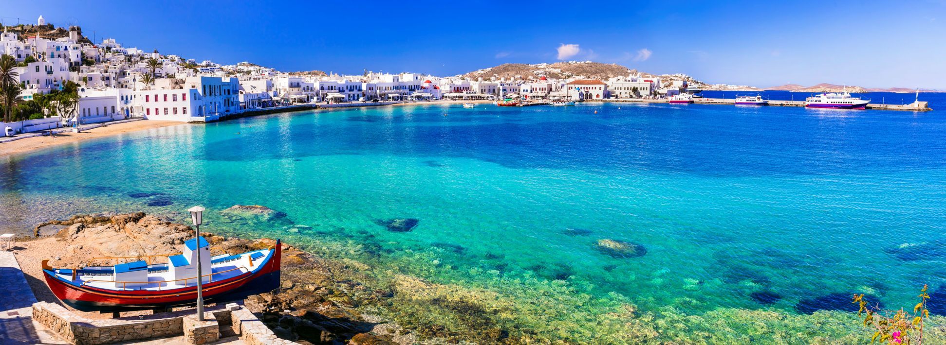 Una cittadina costiera con edifici bianchi si affaccia su una baia di acqua turchese cristallina sotto un cielo azzurro, con una piccola barca sulla riva.