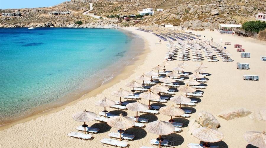 Vista dall'alto di una spiaggia sabbiosa fiancheggiata da file di ombrelloni di paglia e sedie a sdraio accanto a un'acqua limpida e turchese.