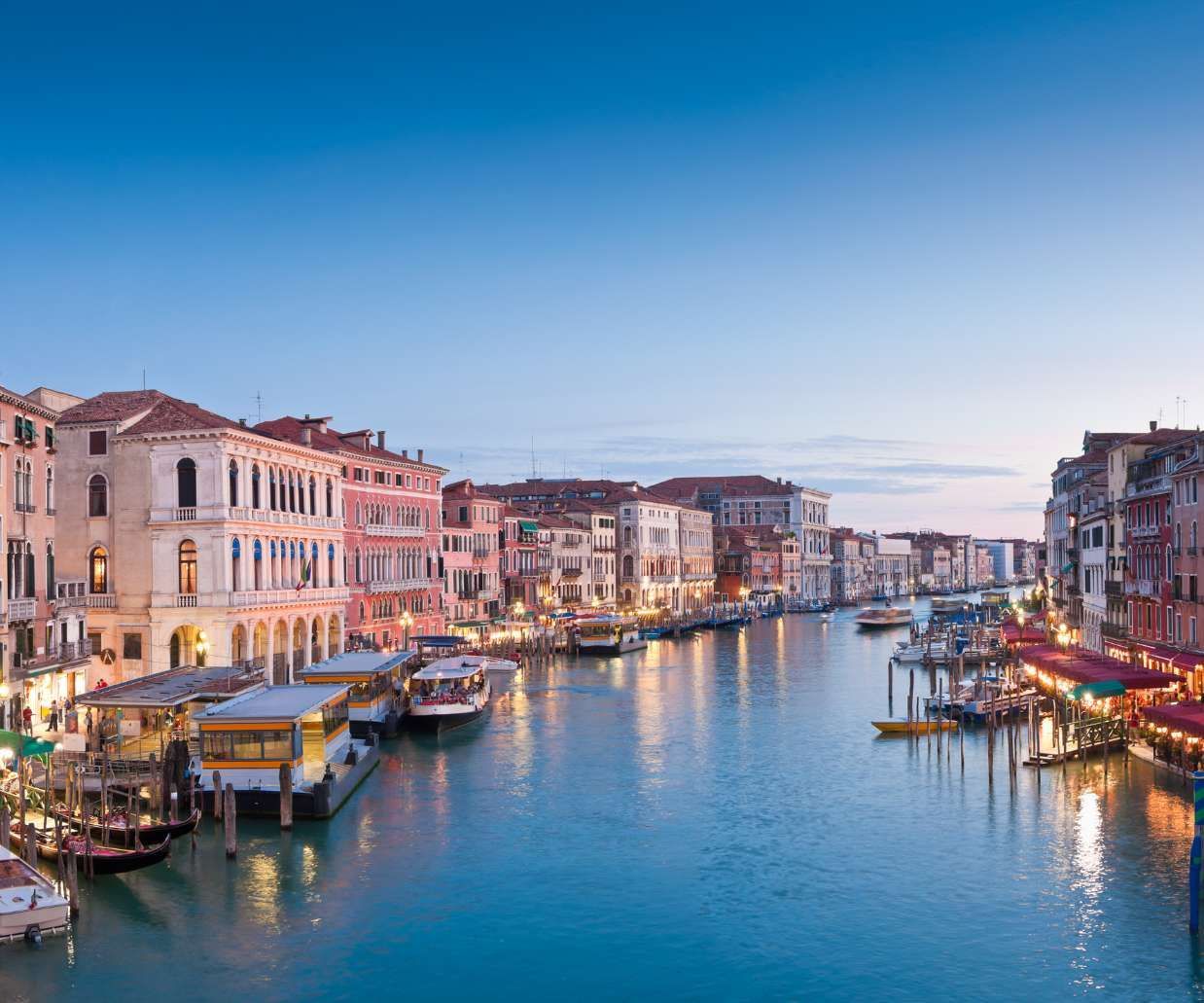 Illuminated historic buildings line a wide canal filled with boats and vaporettos under a clear blue sky at dusk.