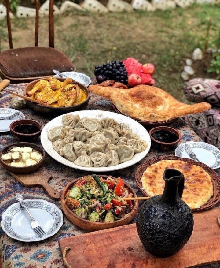 Un tavolo apparecchiato all'aperto con un banchetto di piatti tradizionali georgiani come i khinkali, pane, insalata, stufato e frutta fresca.