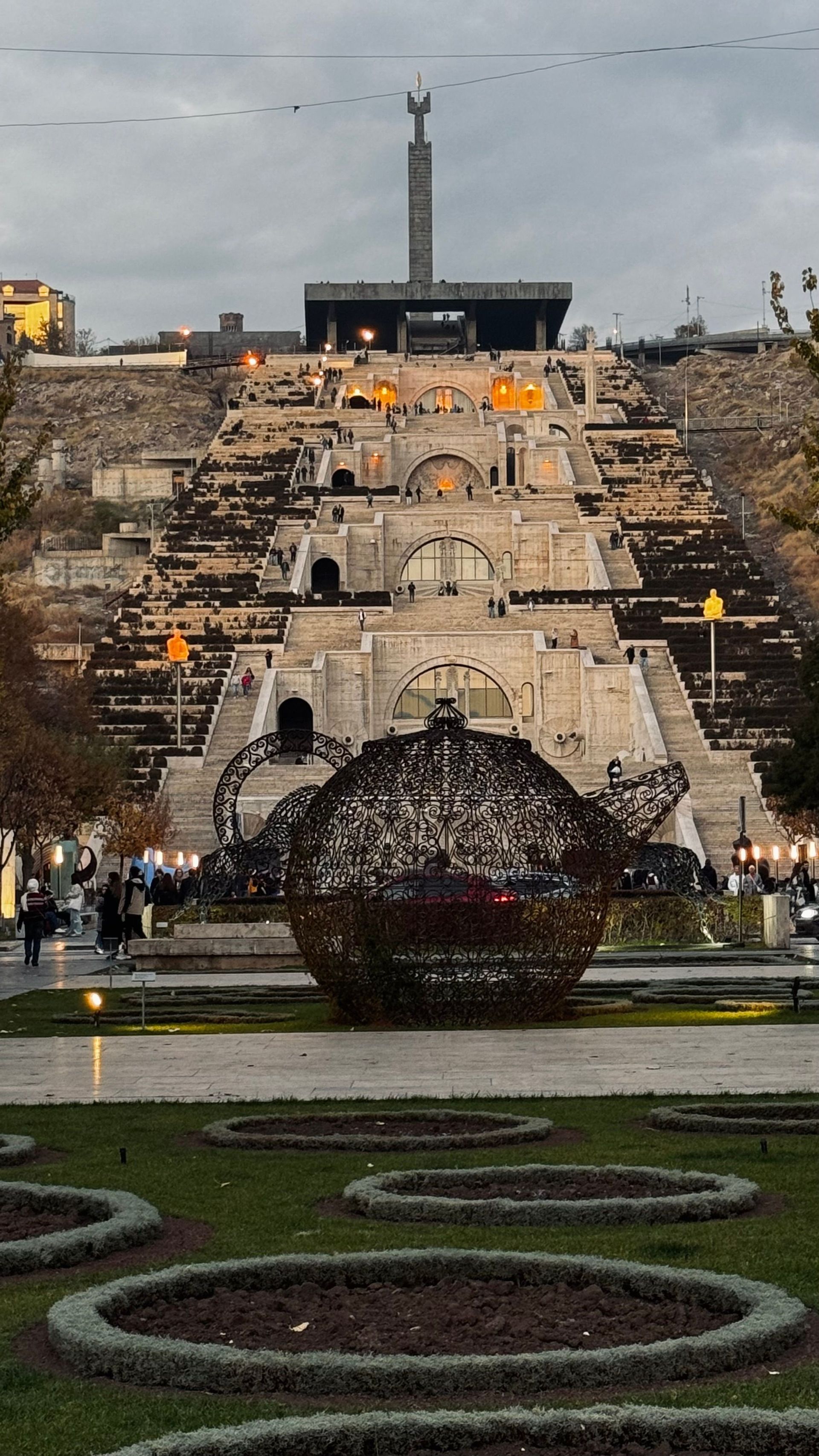 Una grande e intricata scultura in metallo a forma di teiera si trova in un parco, ai piedi di una monumentale scalinata in pietra a più livelli, al crepuscolo.