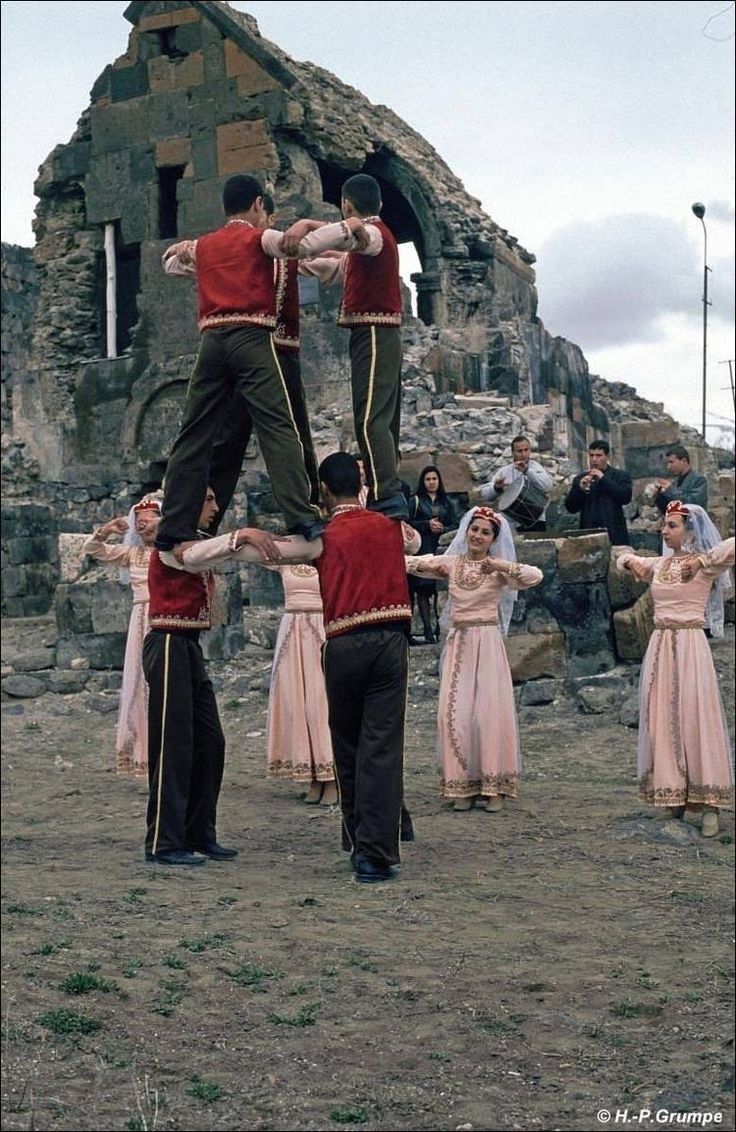 Un gruppo di persone in costumi tradizionali esegue una danza folcloristica, con gli uomini che formano una piramide umana davanti a rovine in pietra.