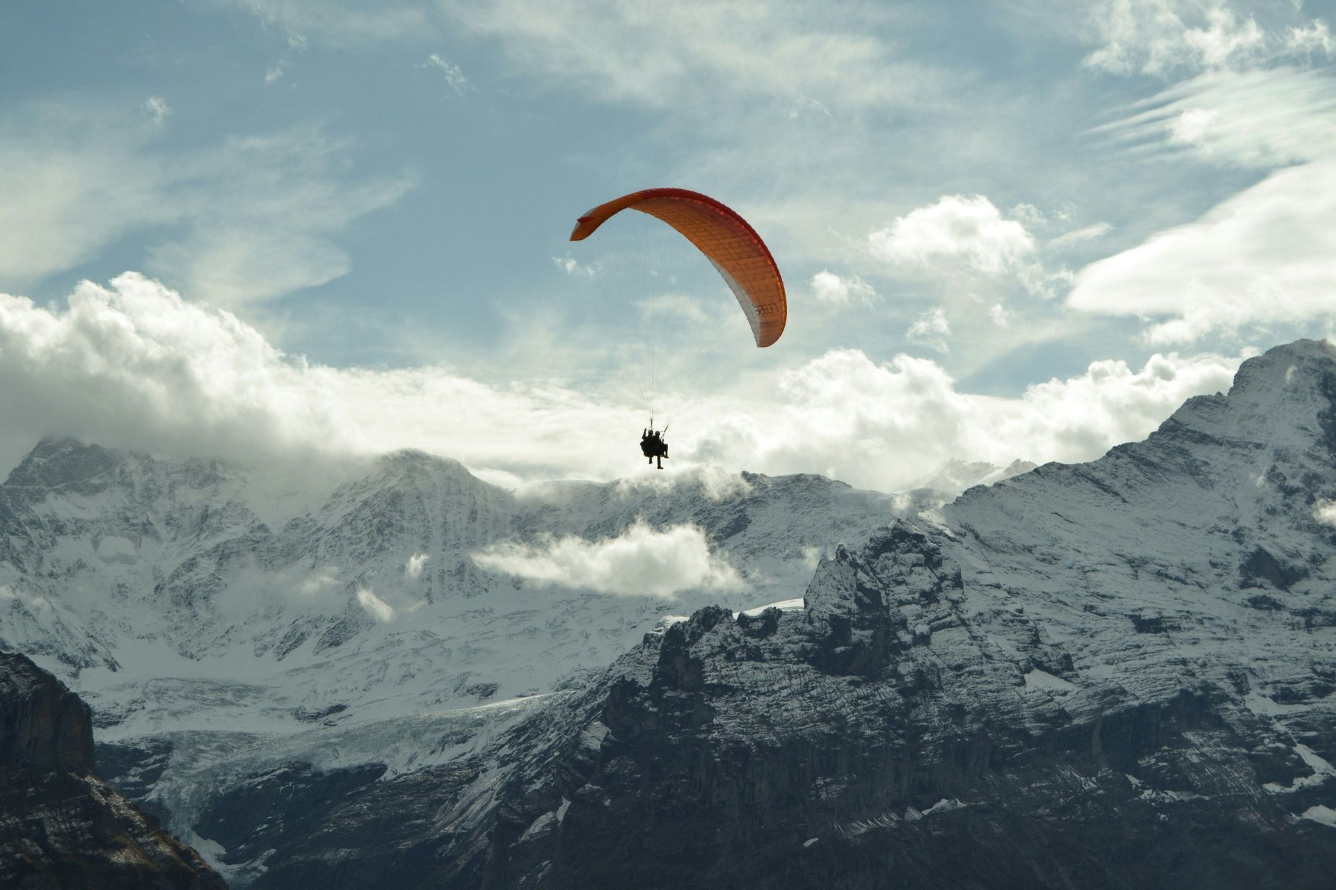 Deux personnes en parapente biplace avec une aile orange s'envolent au-dessus d'une vaste chaîne de montagnes enneigée sous un ciel nuageux.