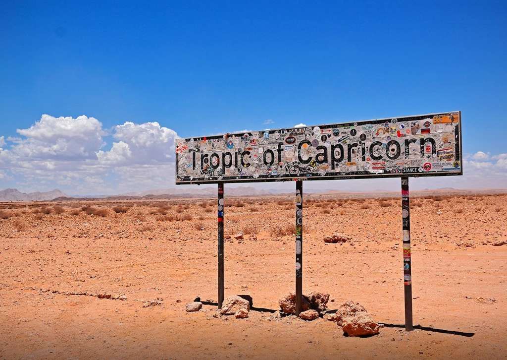 Un grande cartello con la scritta 'Tropico del Capricorno', coperto di adesivi, si erge in un vasto paesaggio desertico sotto un cielo blu.