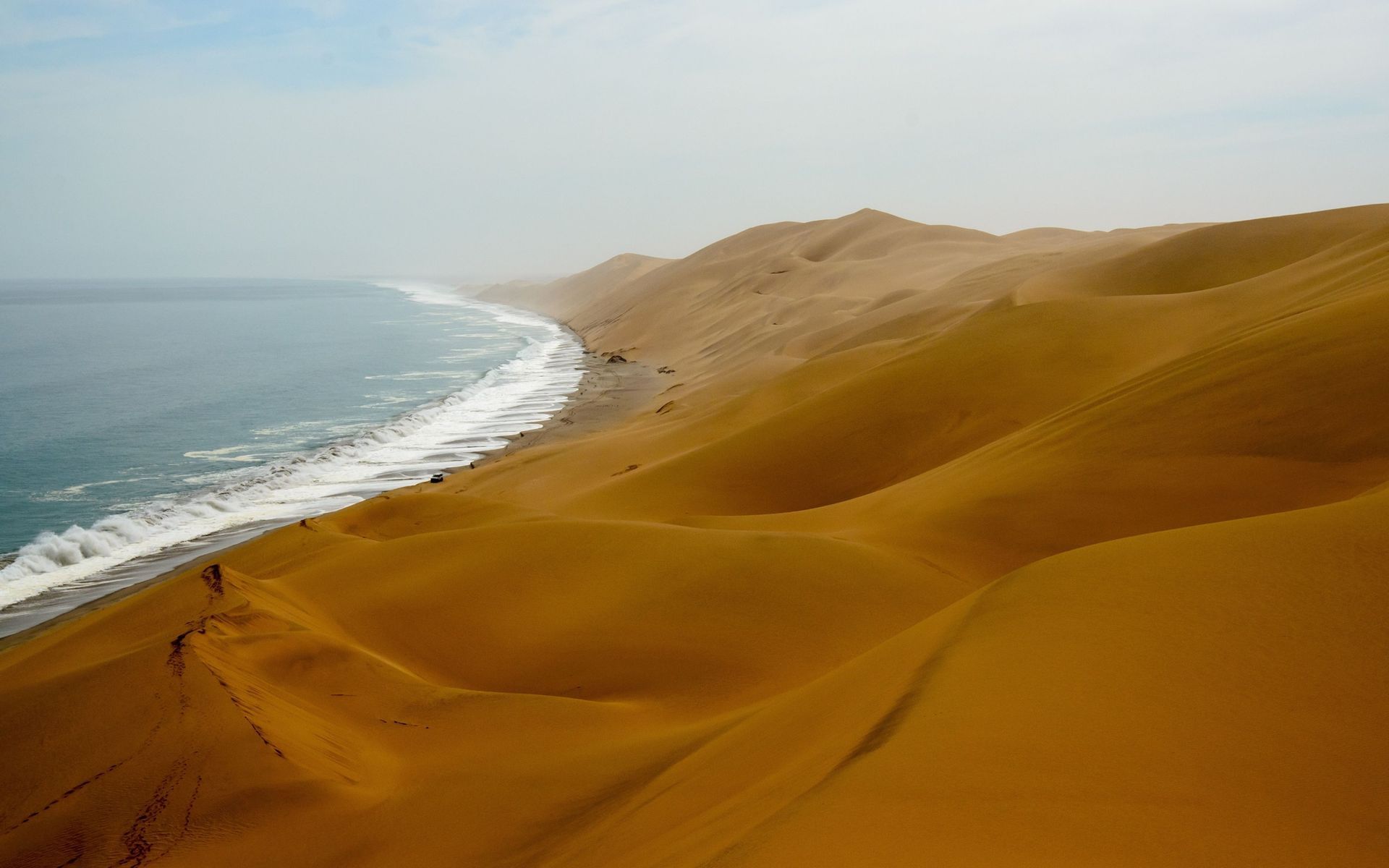 Dune di sabbia dorata si estendono lungo una costa, incontrando l'oceano blu con onde bianche sotto un cielo pallido.