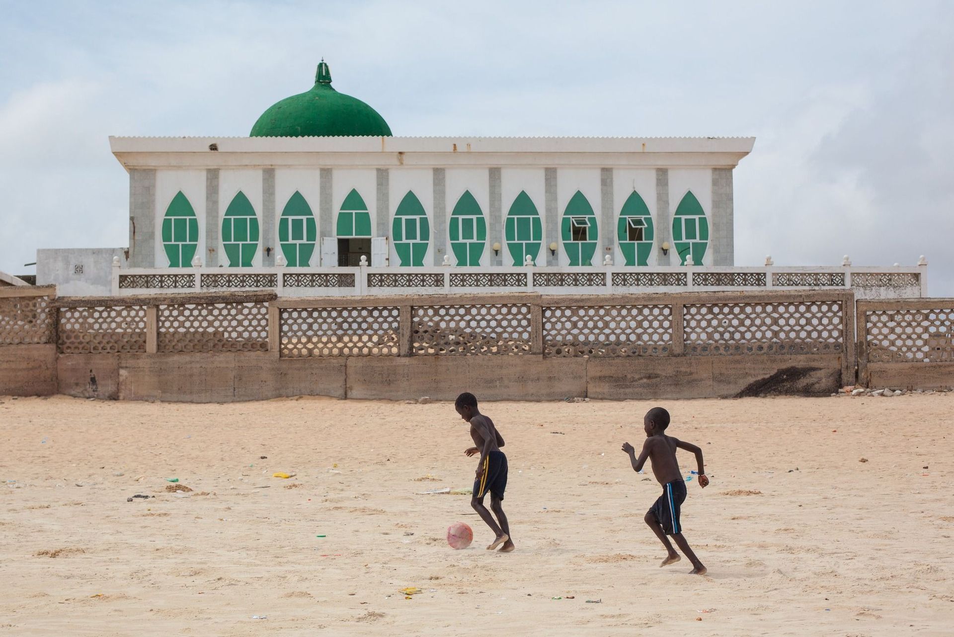 Due giovani ragazzi giocano a calcio su un terreno sabbioso davanti a una grande moschea bianca e verde con una cupola.