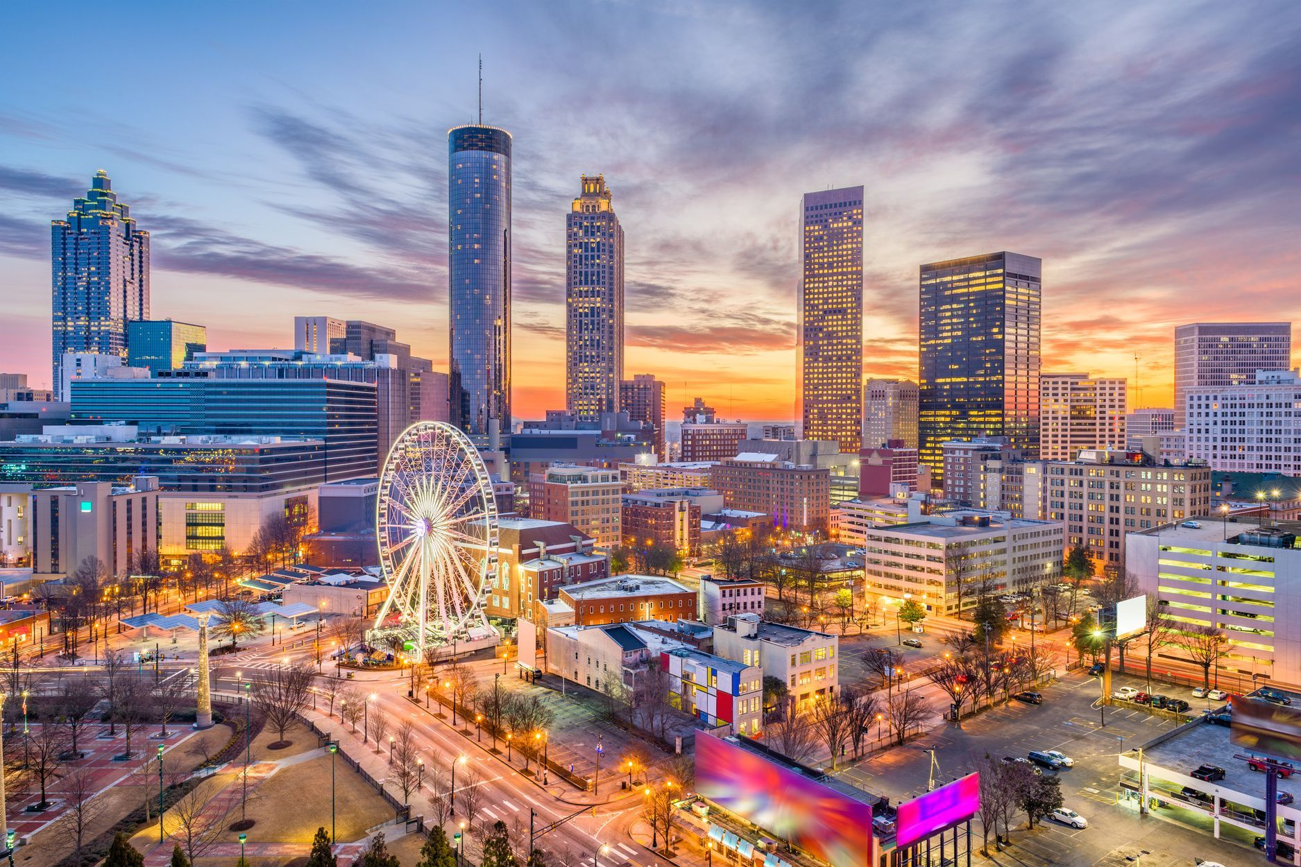 Un panorama urbain avec des gratte-ciel illuminés et une grande roue sous un ciel de coucher de soleil coloré.