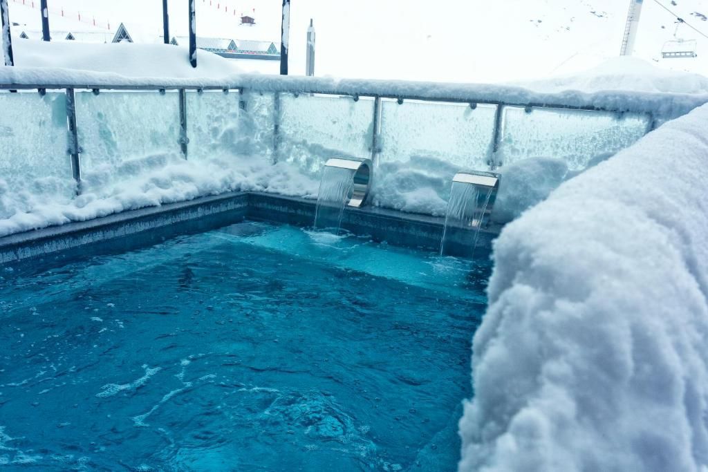 An outdoor hot tub with two waterfall jets flowing into the blue water, surrounded by deep snow at a mountain resort.