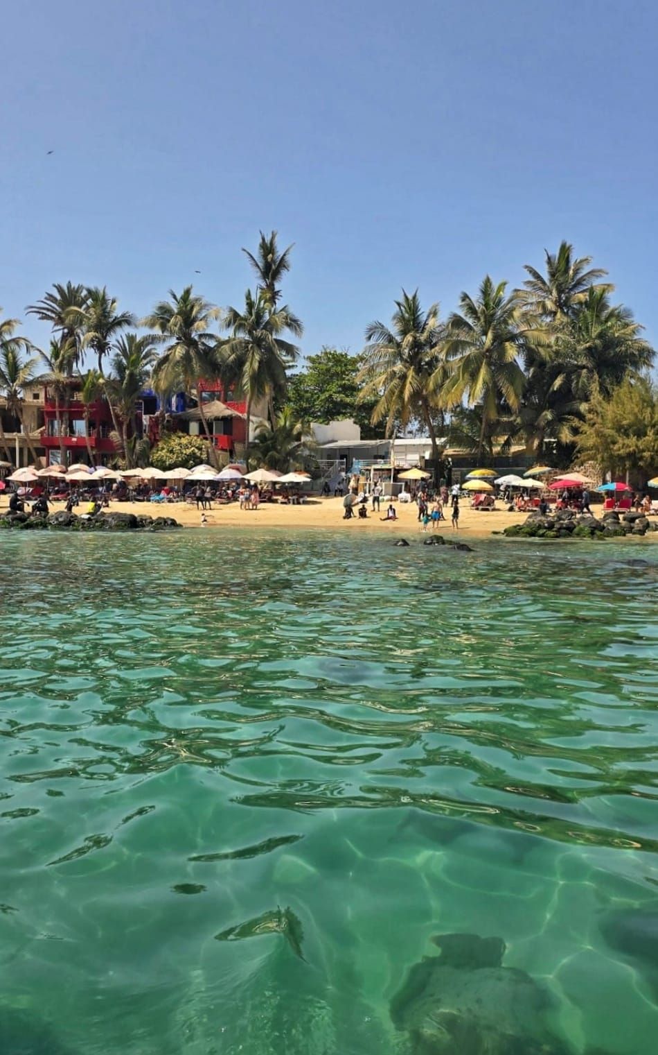 Una vista dall'acqua limpida e turchese di una spiaggia sabbiosa affollata di persone sotto gli ombrelloni, con palme e edifici sullo sfondo.