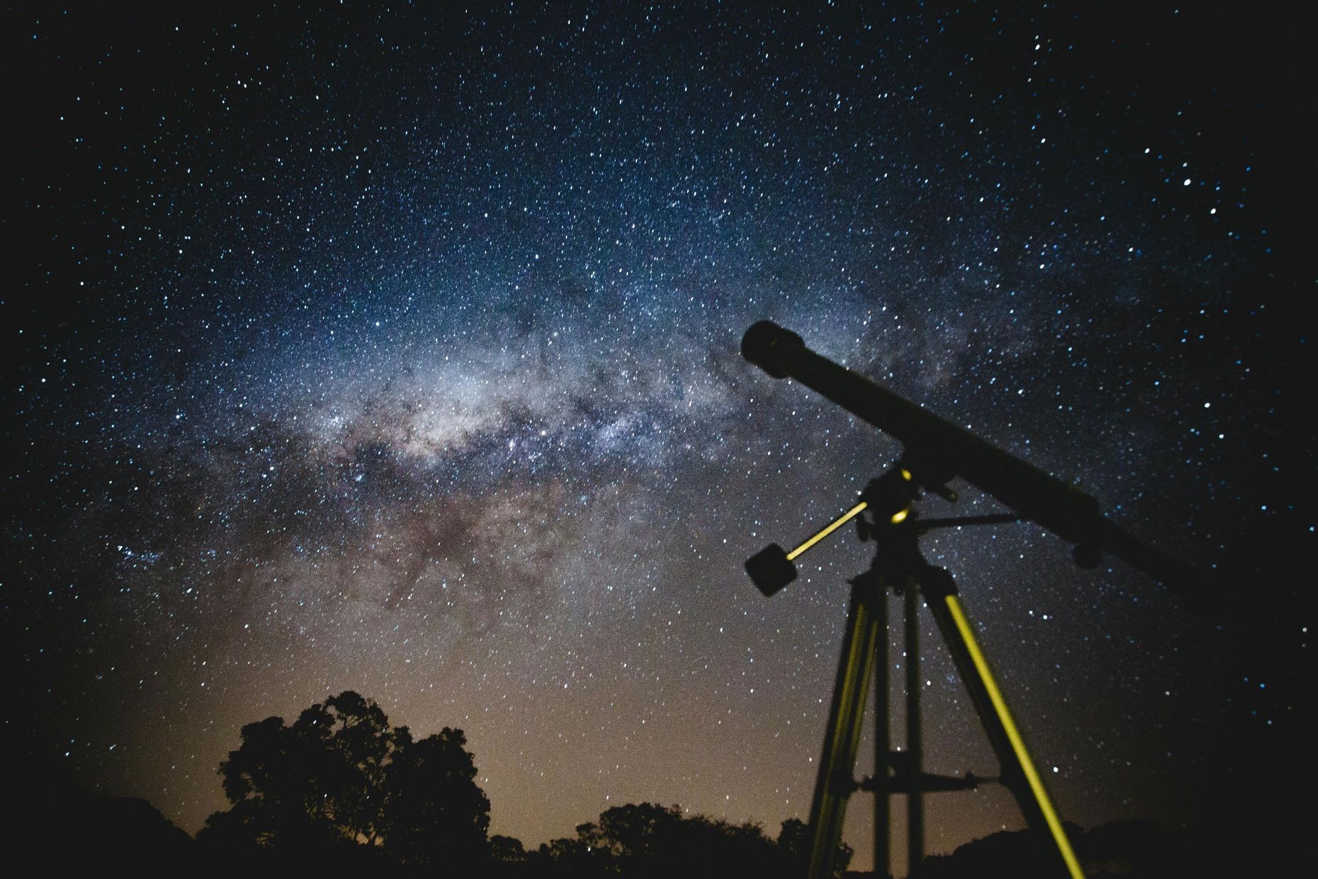 Un telescopio su treppiede si staglia contro un cielo notturno stellato con la Via Lattea sopra la sagoma degli alberi.