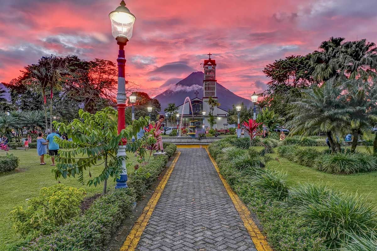Un sentiero lastricato attraverso un parco lussureggiante conduce a una chiesa con un vulcano sullo sfondo sotto un cielo rosato al tramonto.
